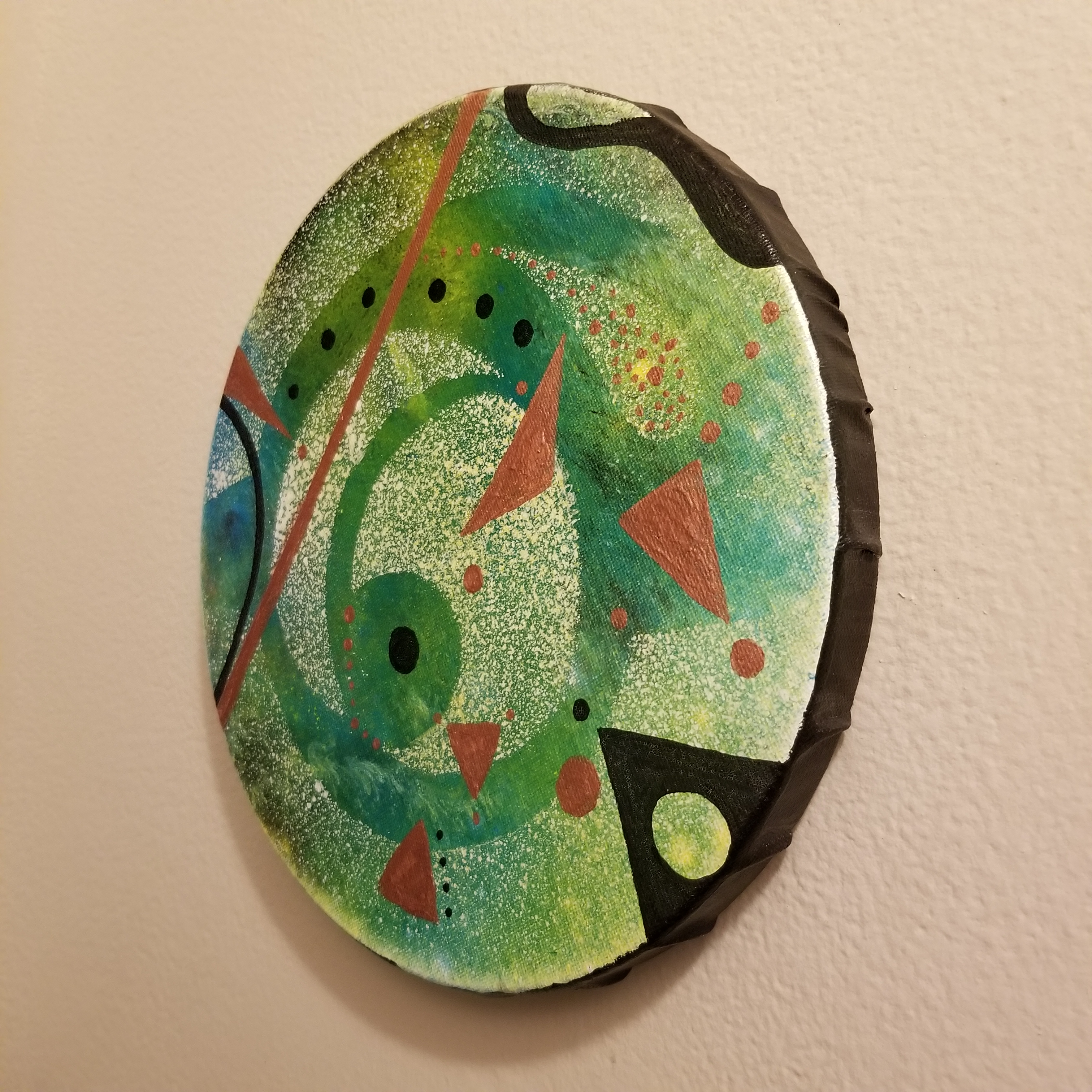 green and metallic copper geometric shapes and lines over a white stencilled spraypainted abstract form andorganic green and yellow color field inspired by pennsylvania dutch hex signs shown at an angle to display the black border of the canvas