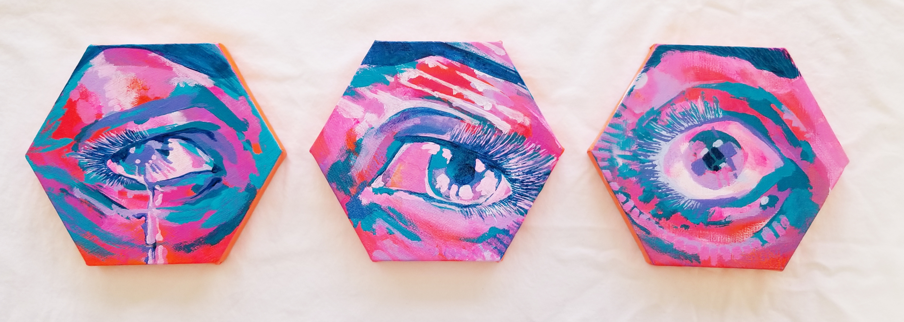 three small hexagonal paintings of single eyes in an abstracted style, painted in bright tropical pink, turquoise blue, pale pink, indigo, and orange